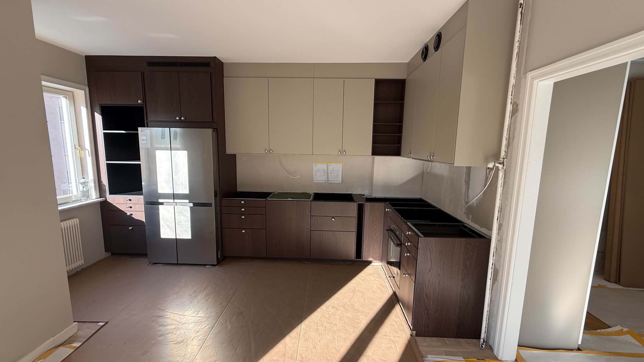 Köksrenovering under montering med mörka ekluckor, beige överskåp och rostfritt kylskåp